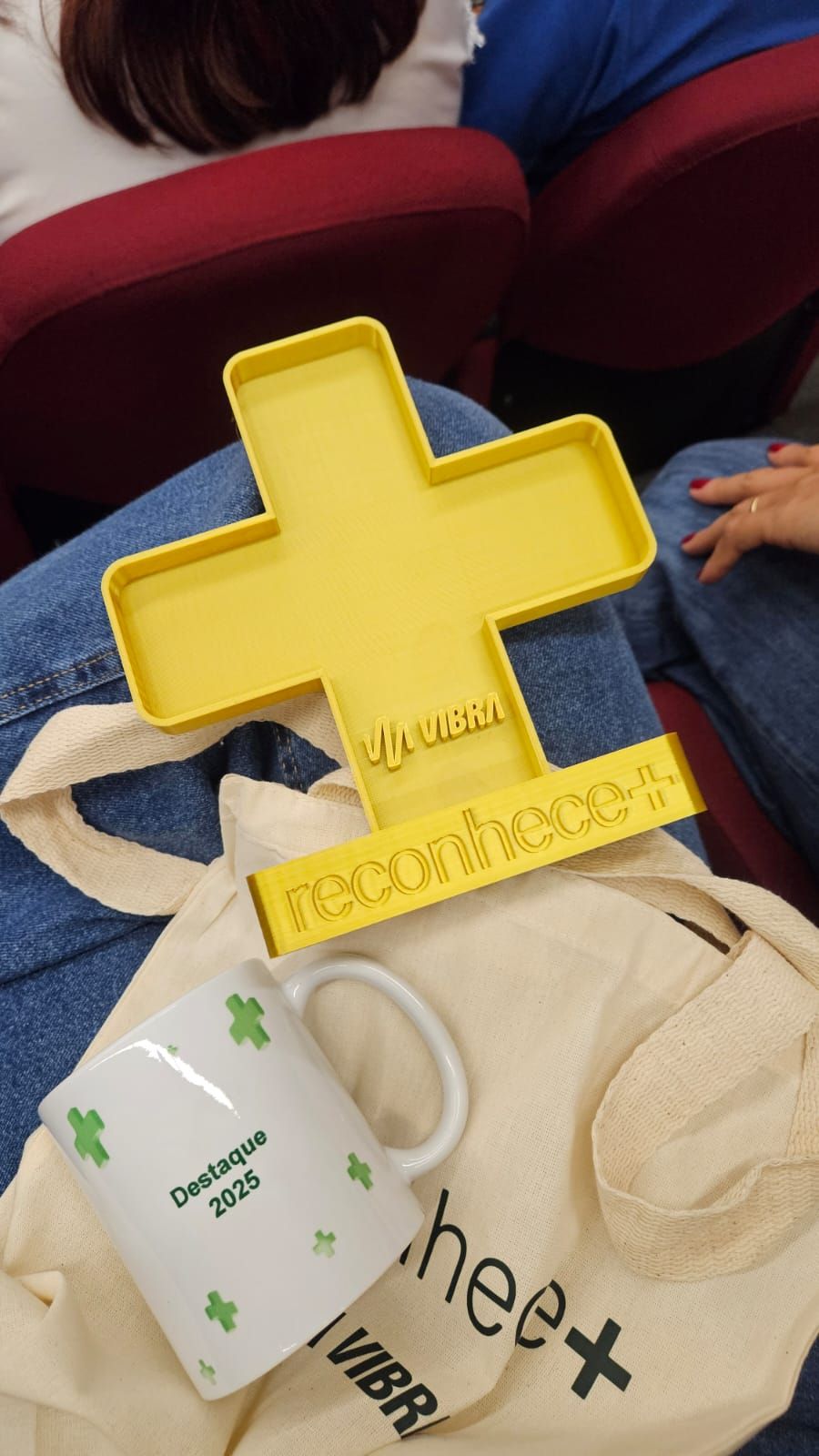 Troféu Reconhece+ em detalhe: formato de + impresso em 3D com logo Vibra em relevo e base com o texto reconhece+, ao lado da caneca Destaque 2025 do kit dos premiados