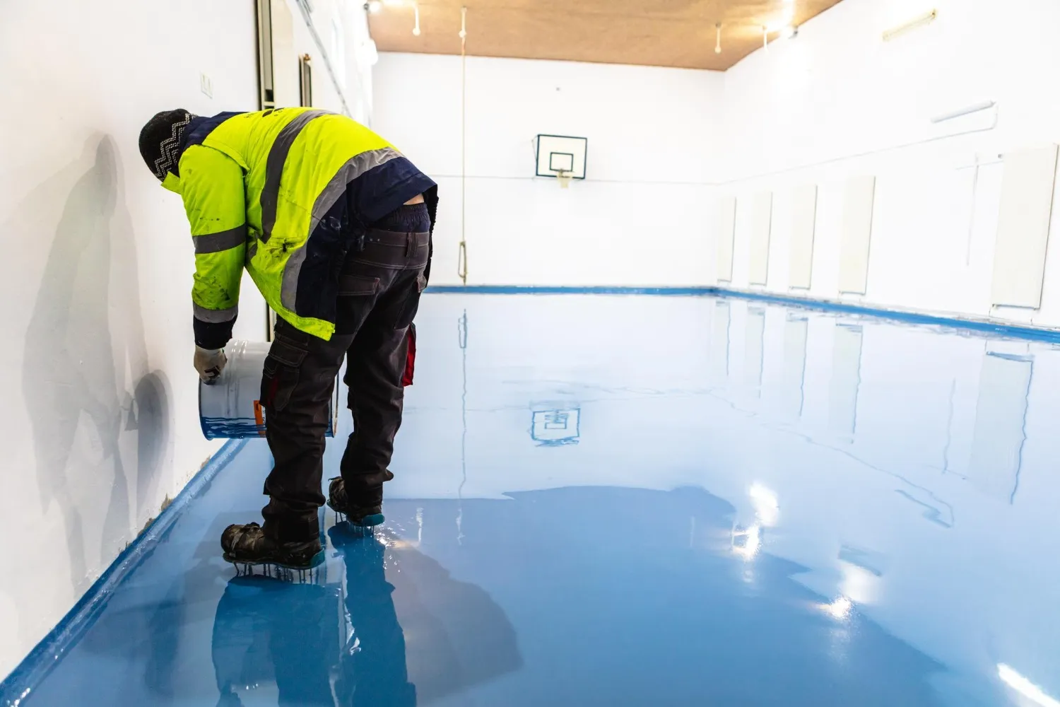 Worker applying blue epoxy resin coating on a large indoor floor, wearing safety gear and specialized spiked shoes, in a gymnasium or sports hall.