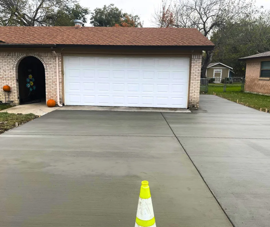 Newly poured and smoothed concrete driveway in front of a residential garage, with visible control joints and a safety cone placed on the surface.