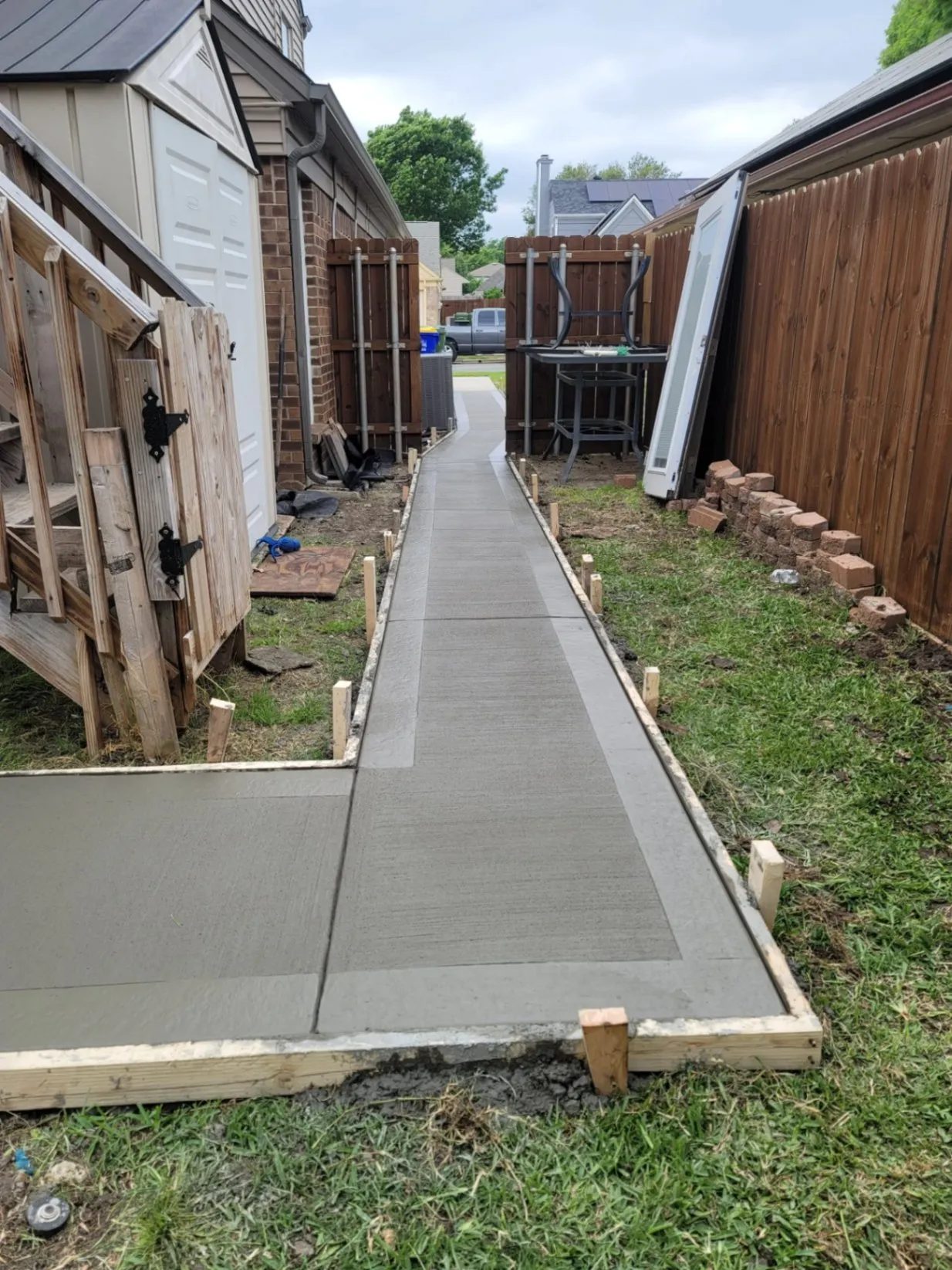 Freshly poured and finished concrete walkway alongside a house, with wooden formwork in place and smooth surface sections leading toward the backyard gate.