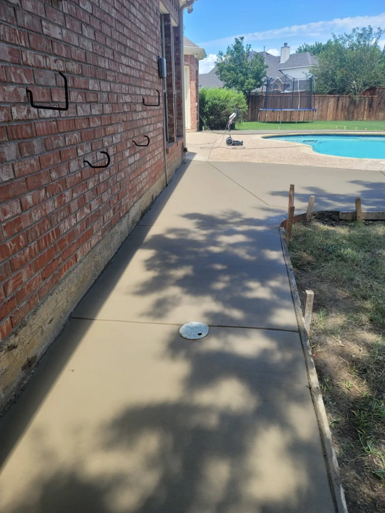 Freshly poured concrete walkway along the side of a brick house, leading to a backyard pool area, with smooth finish and control joints visible.