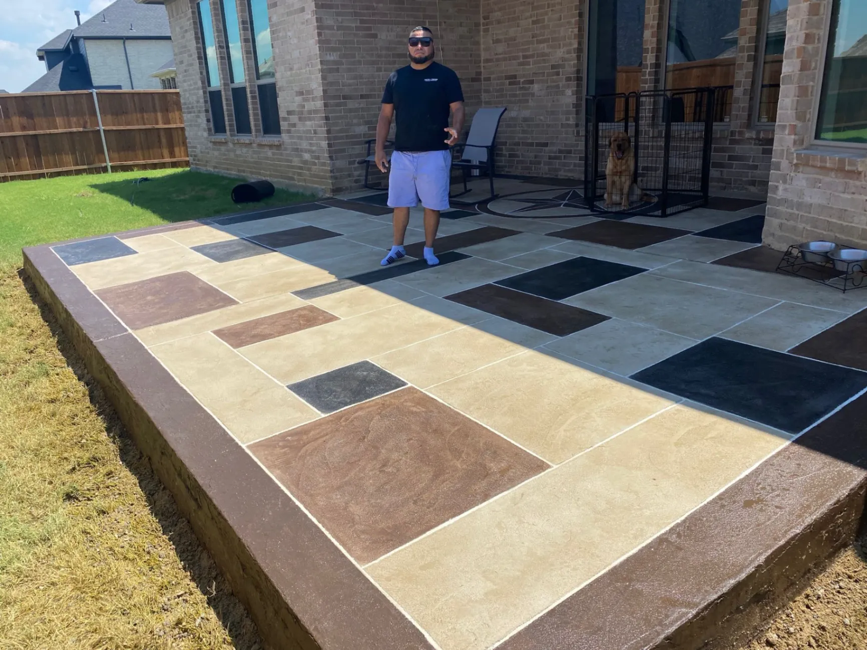 Decorative concrete patio with multicolored stamped and stained square patterns in the backyard of a house, with a man standing on the surface and a dog in the background.