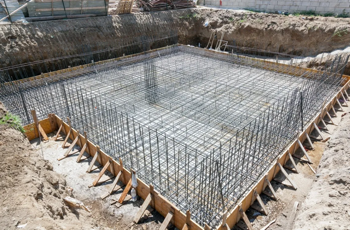 Construction site with a reinforced concrete foundation in progress, showing steel rebar grid arranged within wooden formwork, prepared for pouring concrete.