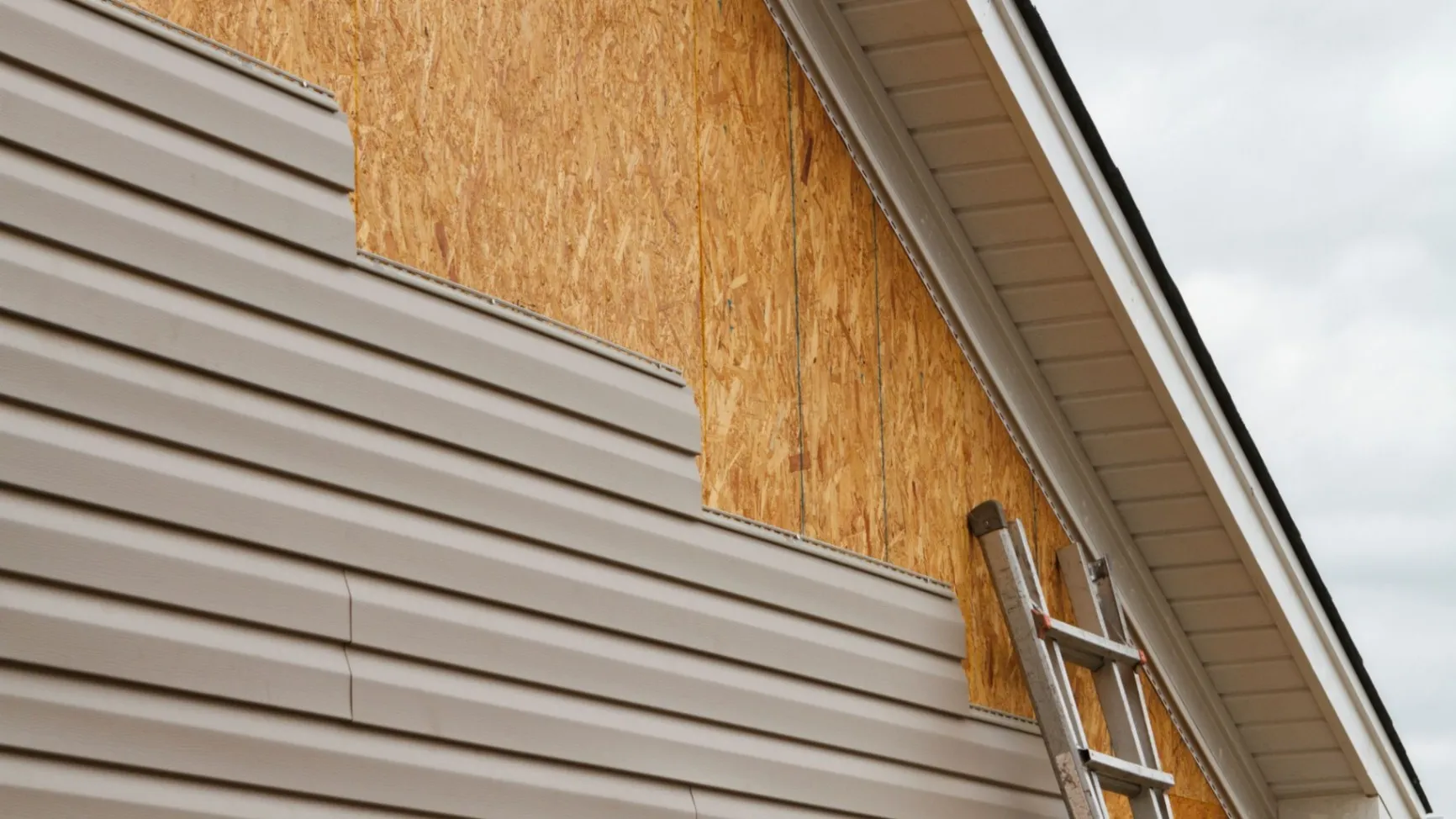 Partially installed beige vinyl siding on a wall with exposed wood sheathing and a ladder.