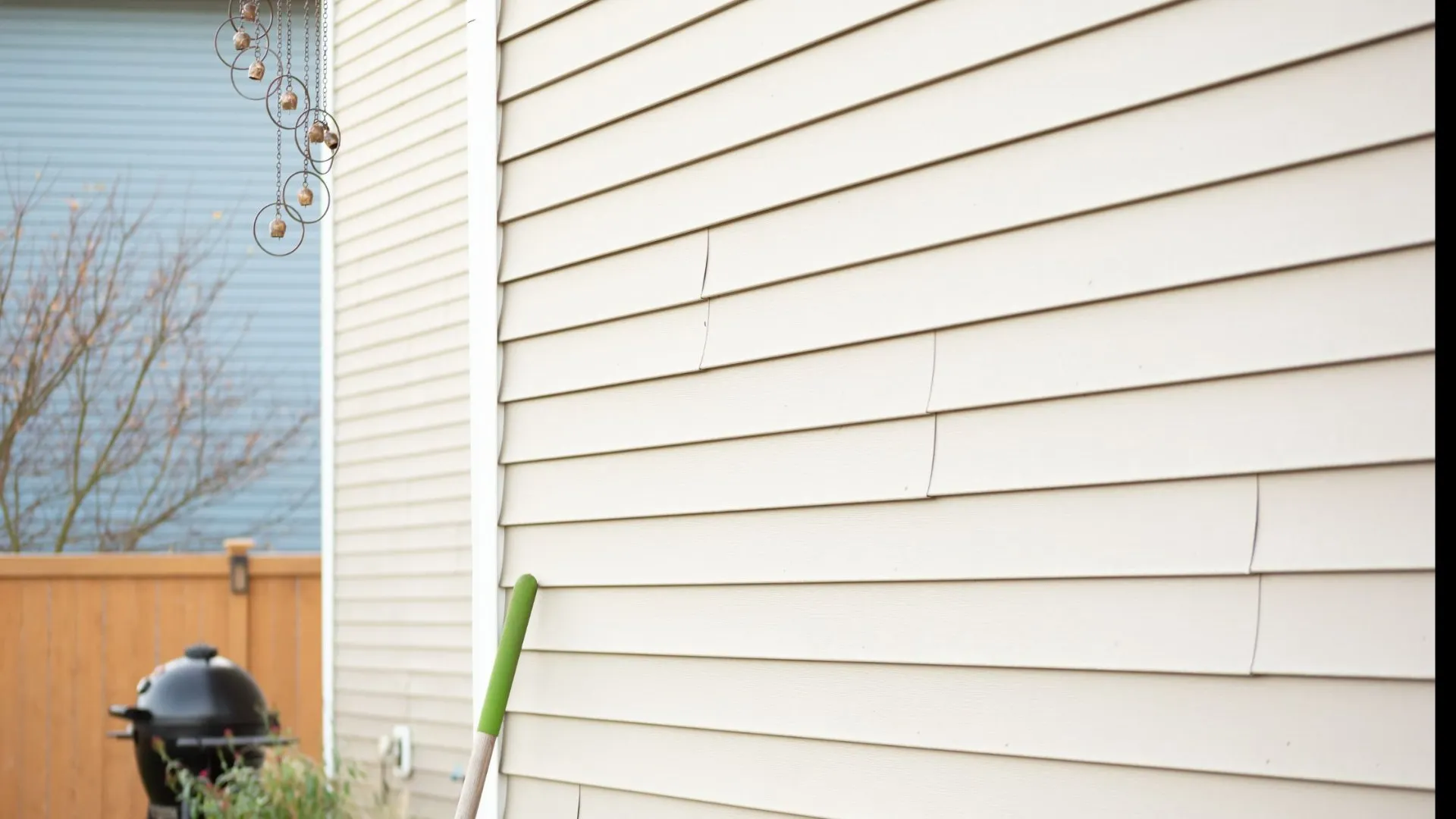 Beige vinyl siding with visible warping and a green-handled tool leaning against the wall.