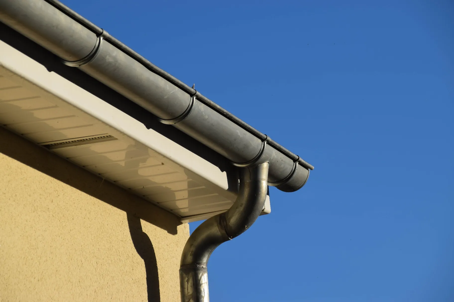 Close-up of a metal gutter system attached to the roof of a beige building against a clear blue sky.