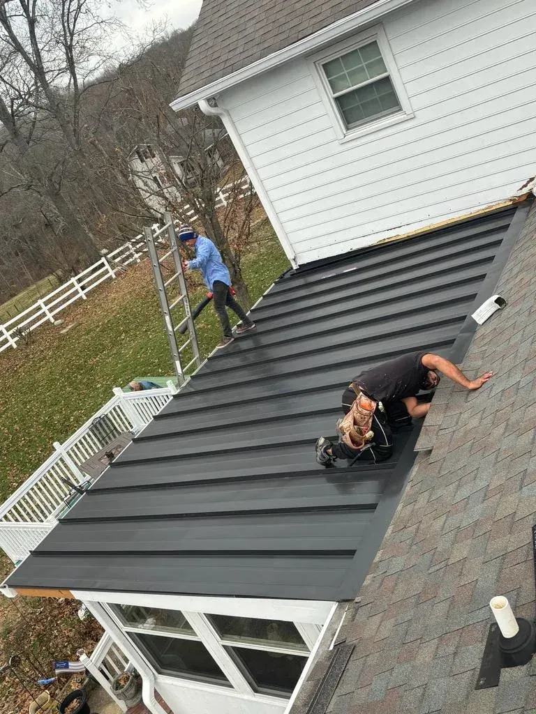 Two workers installing a black metal roof on a house with white siding, using ladders and tools on a cloudy day.