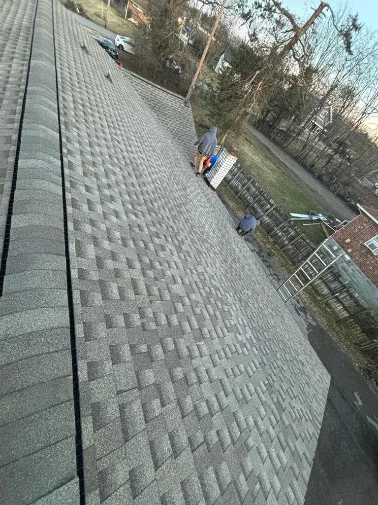 Workers standing on a newly shingled rooftop near a ladder, with trees and houses in the background