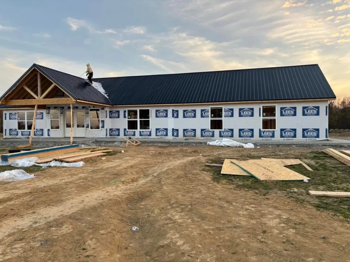 A large single-story house under construction, with a worker standing on the roof, installing a black metal roof.