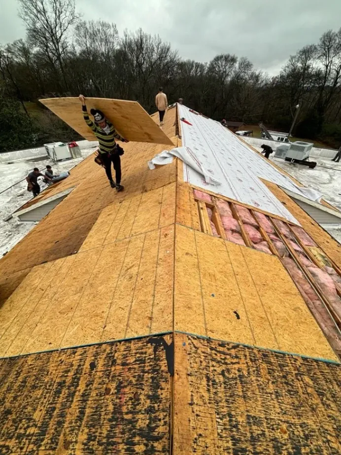 Worker carrying plywood on a roof under construction, with others in the background laying insulation and underlayment.