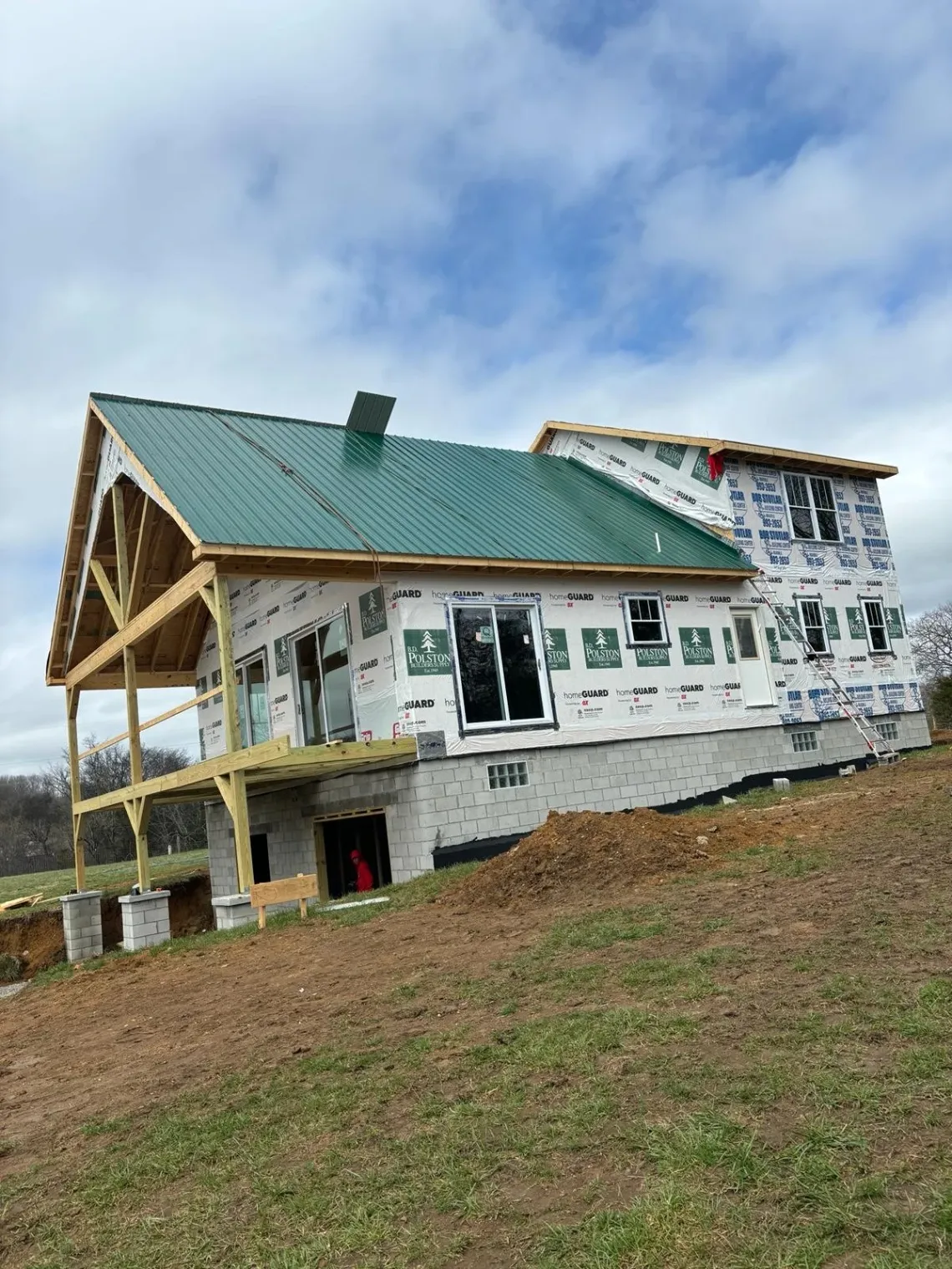 A two-story house under construction with a green metal roof and a wooden deck in progress.