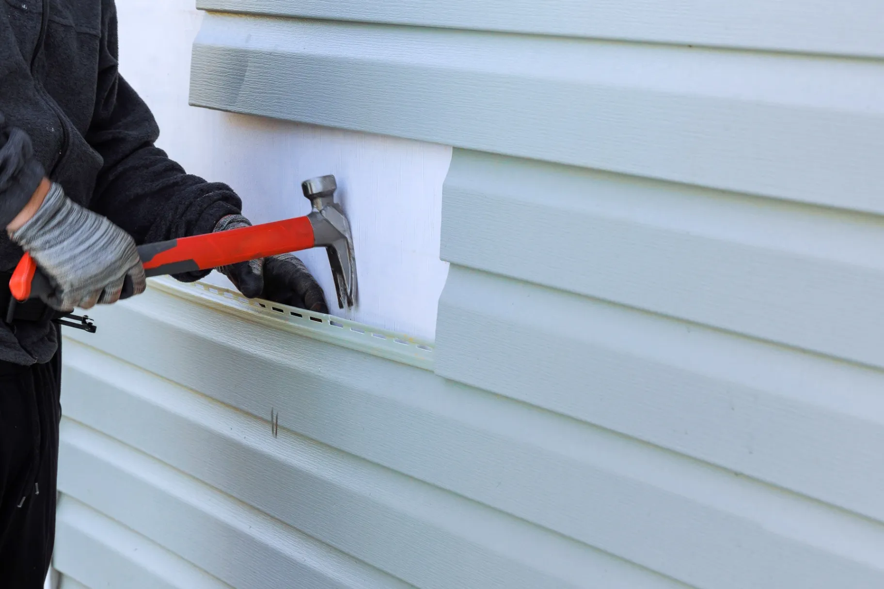 Worker installing vinyl siding with a hammer, aligning panels over a white underlayer.