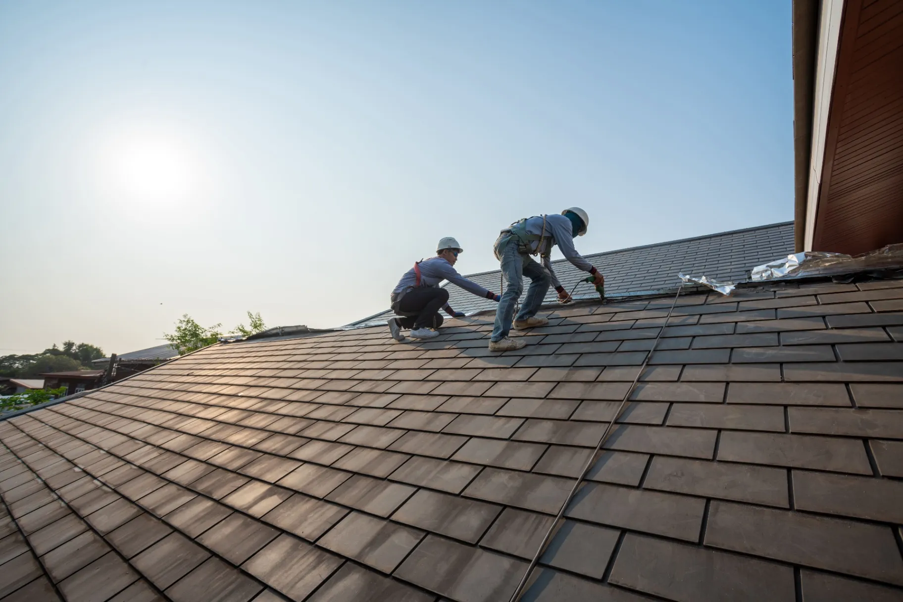 Two roofers wearing safety gear installing tiles on a sloped roof under bright sunlight