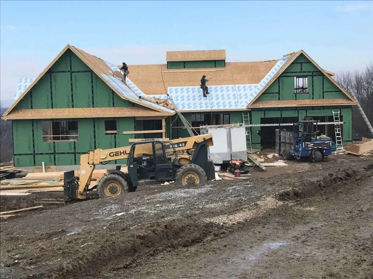 Construction workers installing roofing material on a large green-framed house under construction, with heavy equipment and materials on a muddy site.