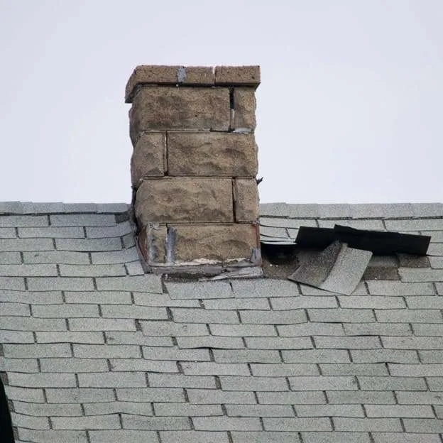 Damaged roof with missing shingles around a brick chimney.