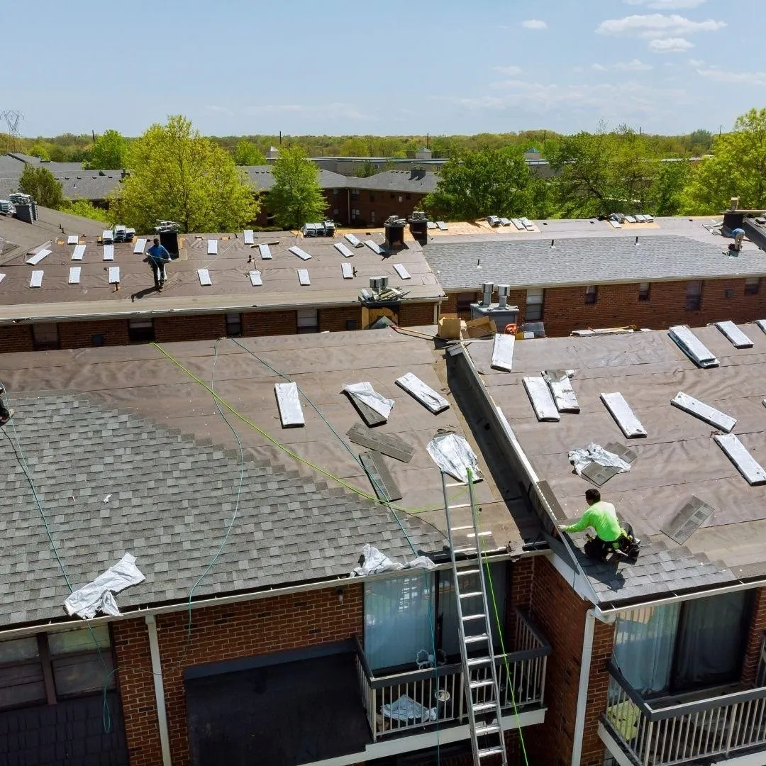 Workers installing new shingles on a large apartment building roof, with roofing materials and tools spread across the surface.