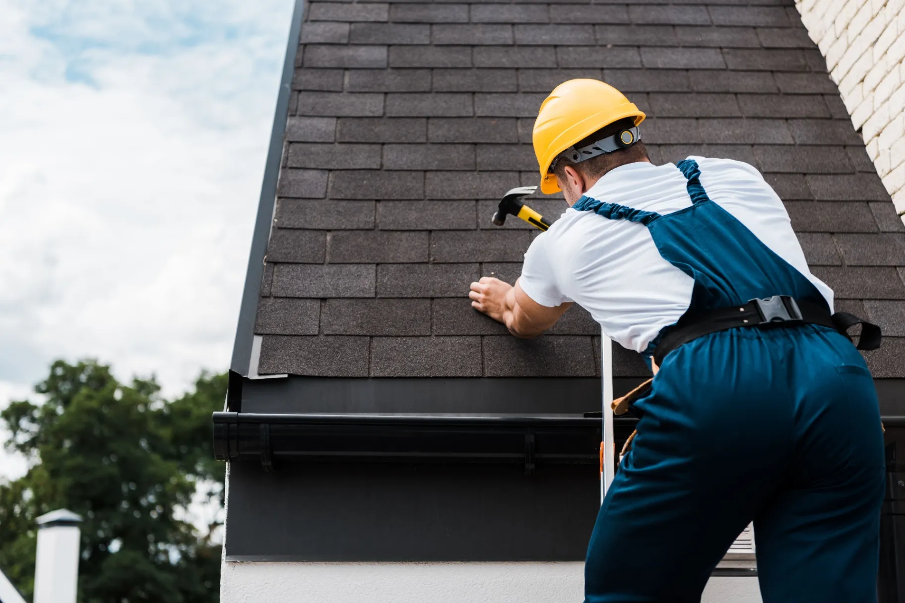 Worker in uniform and helmet installing asphalt shingles on a sloped roof with a hammer."