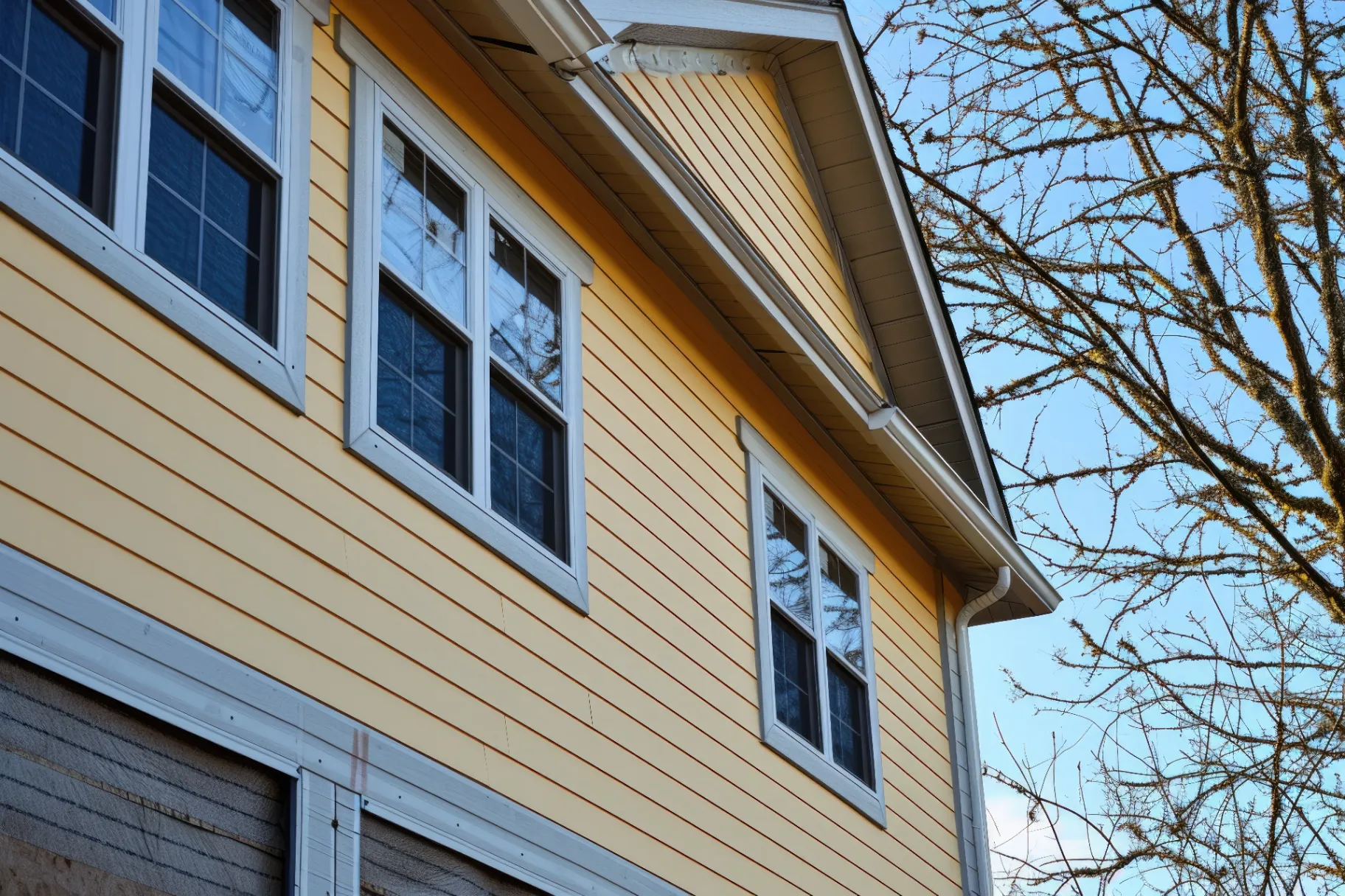 Close-up of a house with freshly installed yellow siding and white-framed windows, set against a clear blue sky and bare tree branches