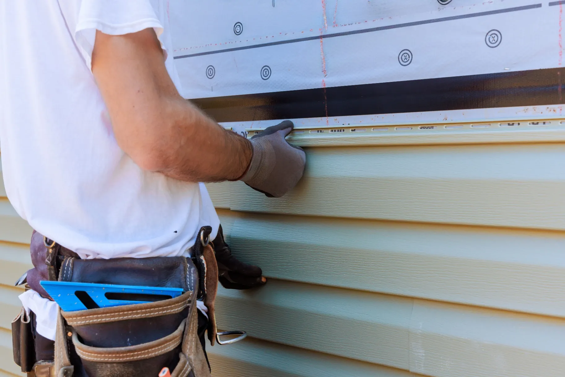 Worker in gloves aligning and installing vinyl siding on a house wall, with tools in a belt.