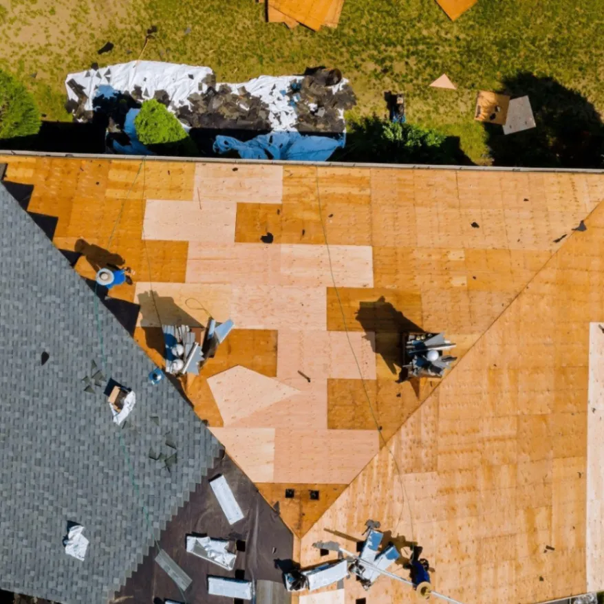 Aerial view of roof replacement in progress, showing plywood decking and partially installed shingles with workers and tools on site.