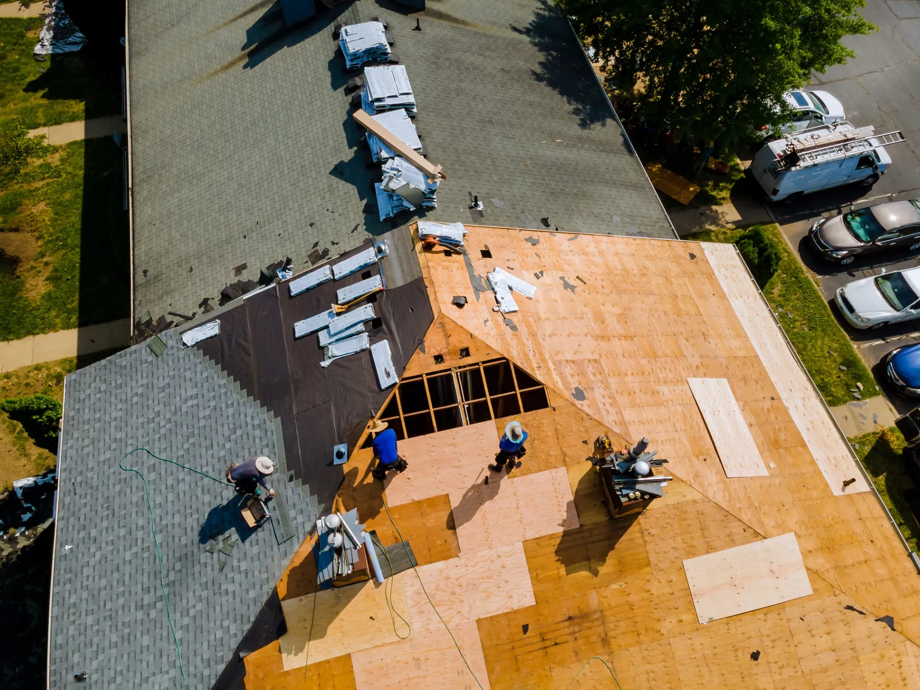 Workers replacing a roof with new shingles, showing partially exposed plywood and stacks of materials.