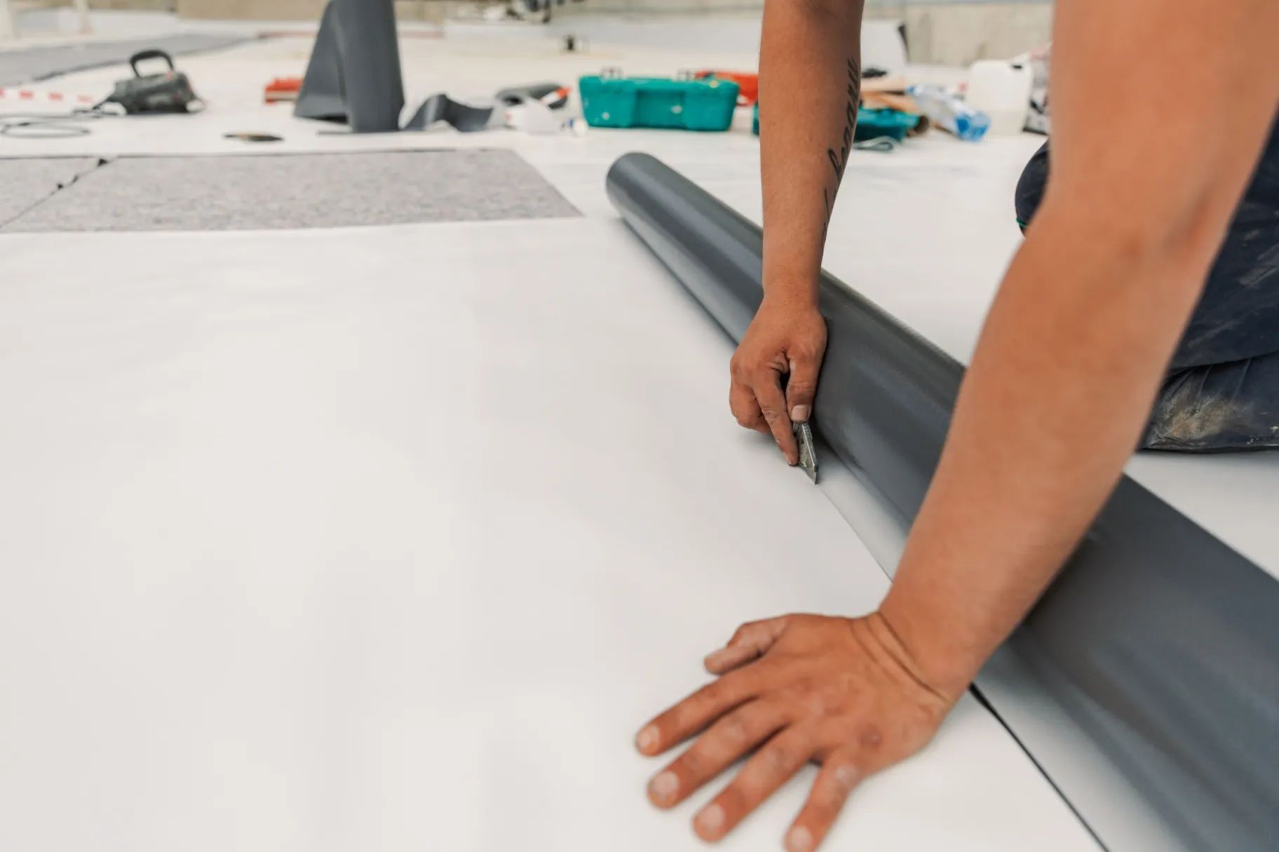 Worker using a utility knife to cut a PVC membrane for roof installation on a flat surface.