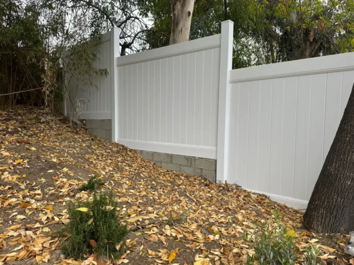 White vinyl privacy fence installed on a sloped yard with block retaining base and surrounding trees