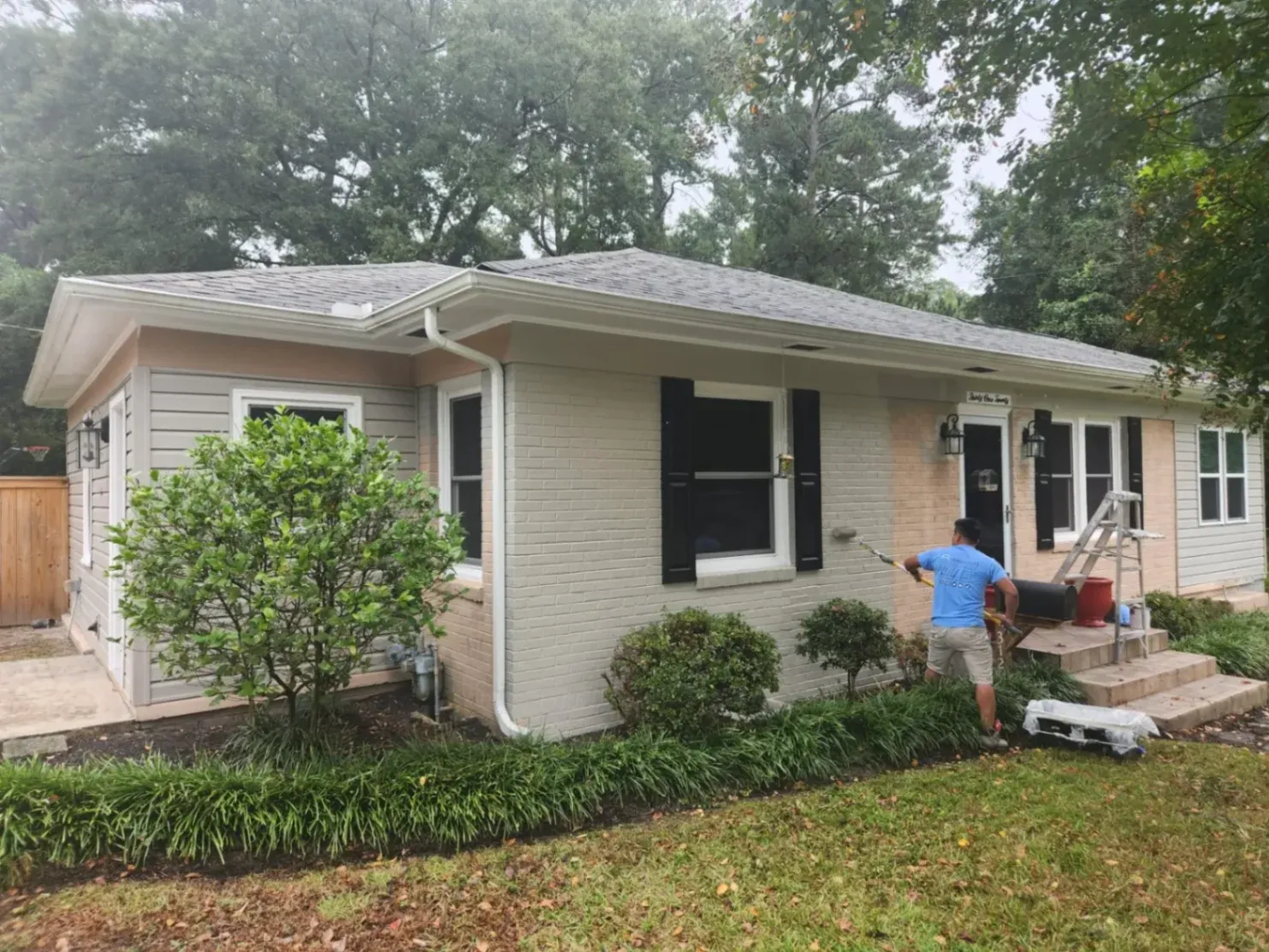 A professional painter applies fresh paint to the brick exterior of a single-story home using a roller, highlighting an exterior painting & renovation service in progress with visible prep materials and a ladder nearby.