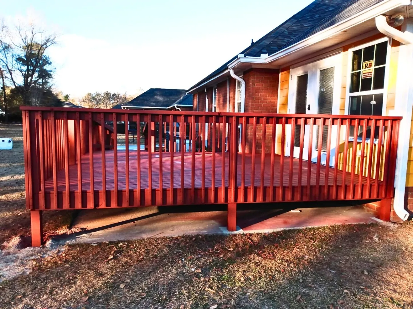 A freshly painted red wooden deck attached to a brick and siding house, featuring vertical railing slats and elevated on support posts above a concrete slab.
