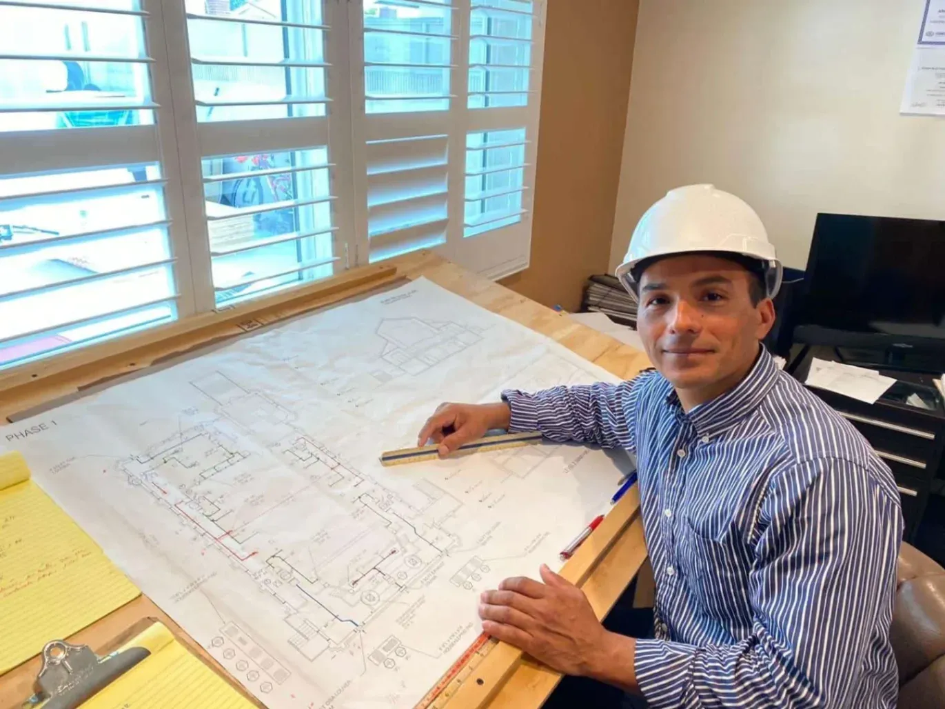 Angel, owner of AAC Heating and Air, is wearing a white hard hat and sits at a drafting table reviewing large building plans, showing the planning and design side of professional HVAC installation work in Garden Grove, CA.