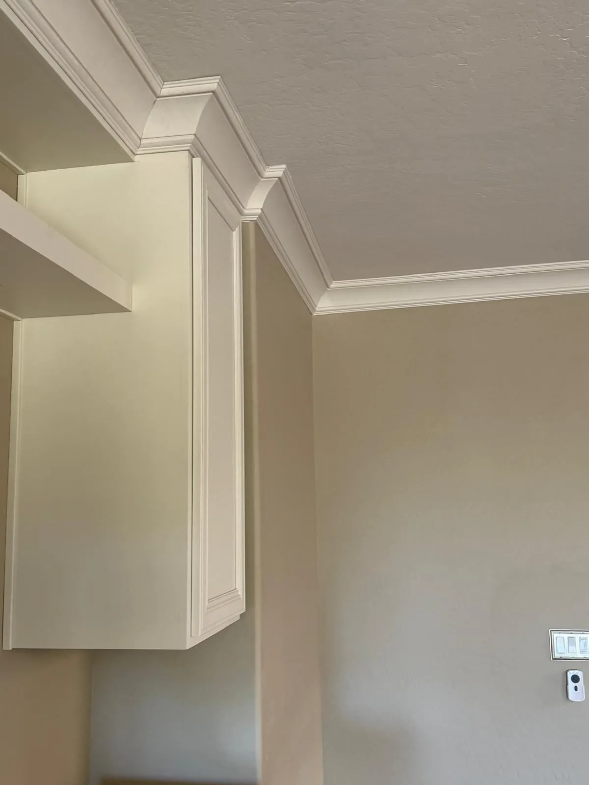 Close-up view of white crown molding installed at the junction of beige walls and a light ceiling, with custom trim continuing seamlessly over a built-in cabinet. The layered design adds a polished, architectural finish to the upper corners of the room.