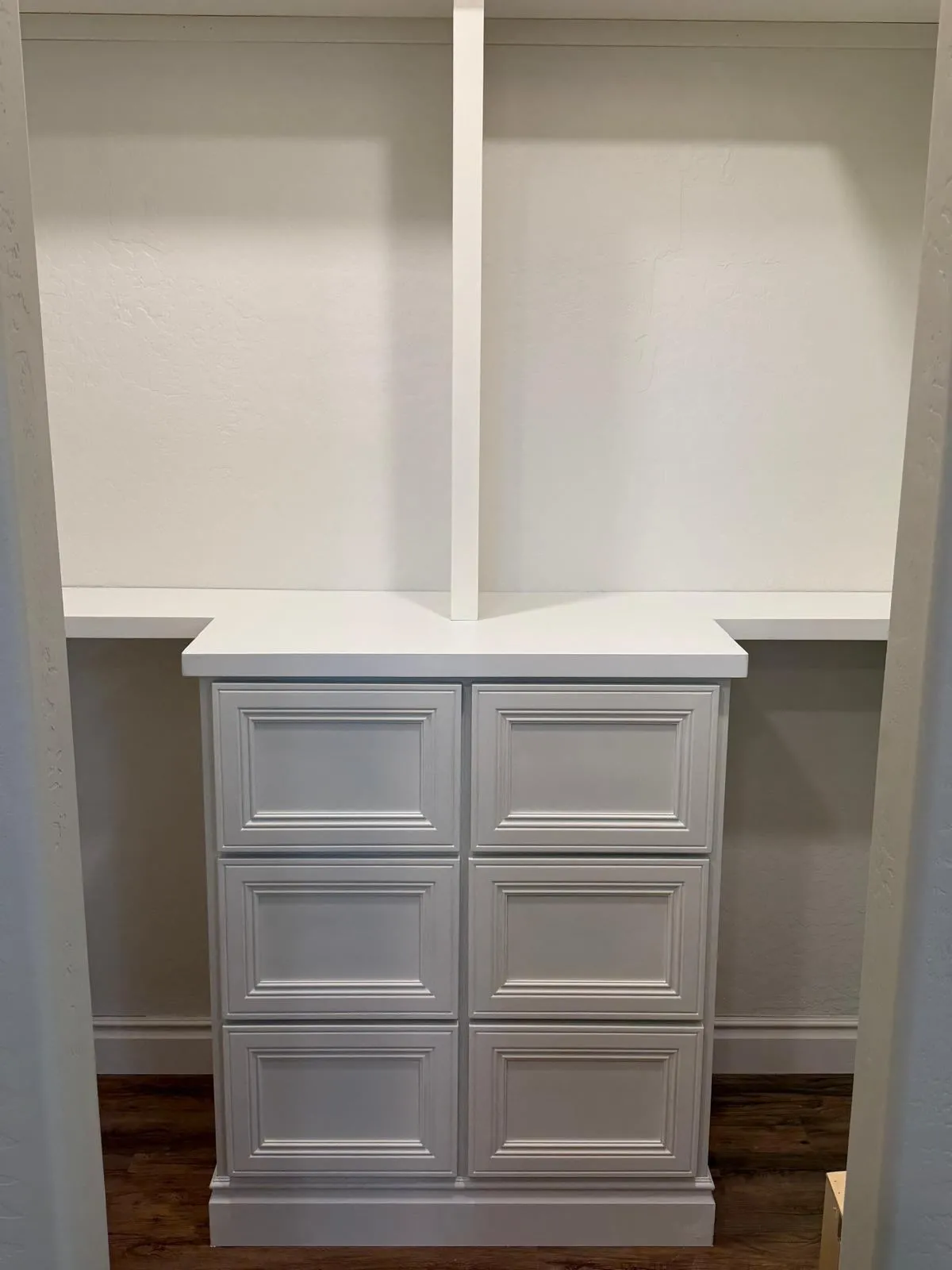 Custom closet installation featuring a white built-in drawer unit with six paneled fronts and a smooth countertop surface. Surrounding shelving and vertical dividers provide additional open storage, all set against a white wall with dark wood flooring below.