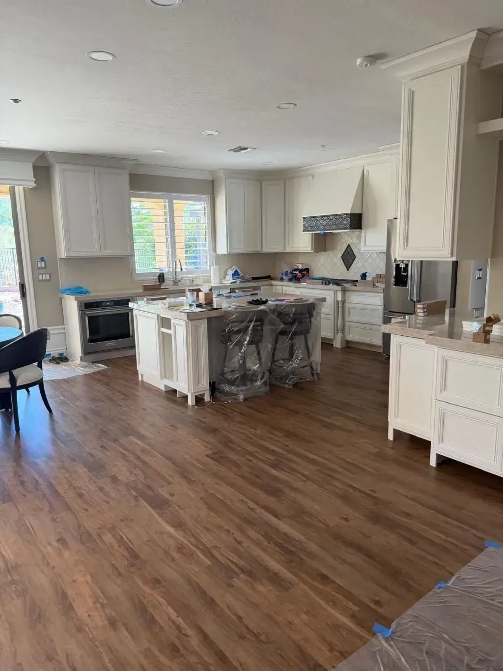 Open-concept kitchen and dining area featuring LVP or engineered wood flooring in a rich, warm tone that complements the cream-colored cabinetry. The space includes a central island with barstools still wrapped in protective plastic, granite countertops, stainless steel appliances, and recessed lighting for a clean, modern finish.