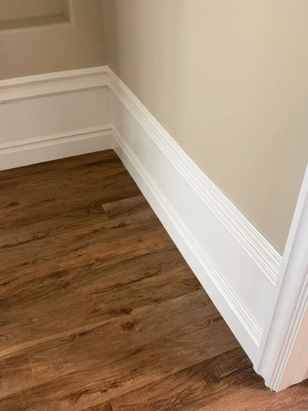 Corner of a room with tall white baseboards featuring layered molding detail along beige walls. The floor is covered in medium-tone wood planks with a natural grain pattern.