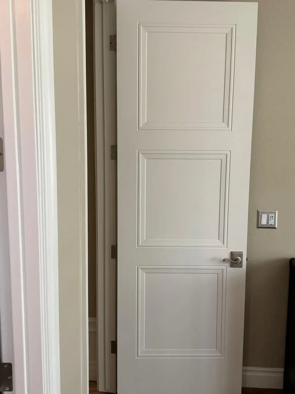 Interior door replacement featuring a white, three-panel door with raised trim detailing and a brushed nickel lever handle. The door is partially open, framed by beige walls and white casing, showcasing a clean, updated look in a modern home.