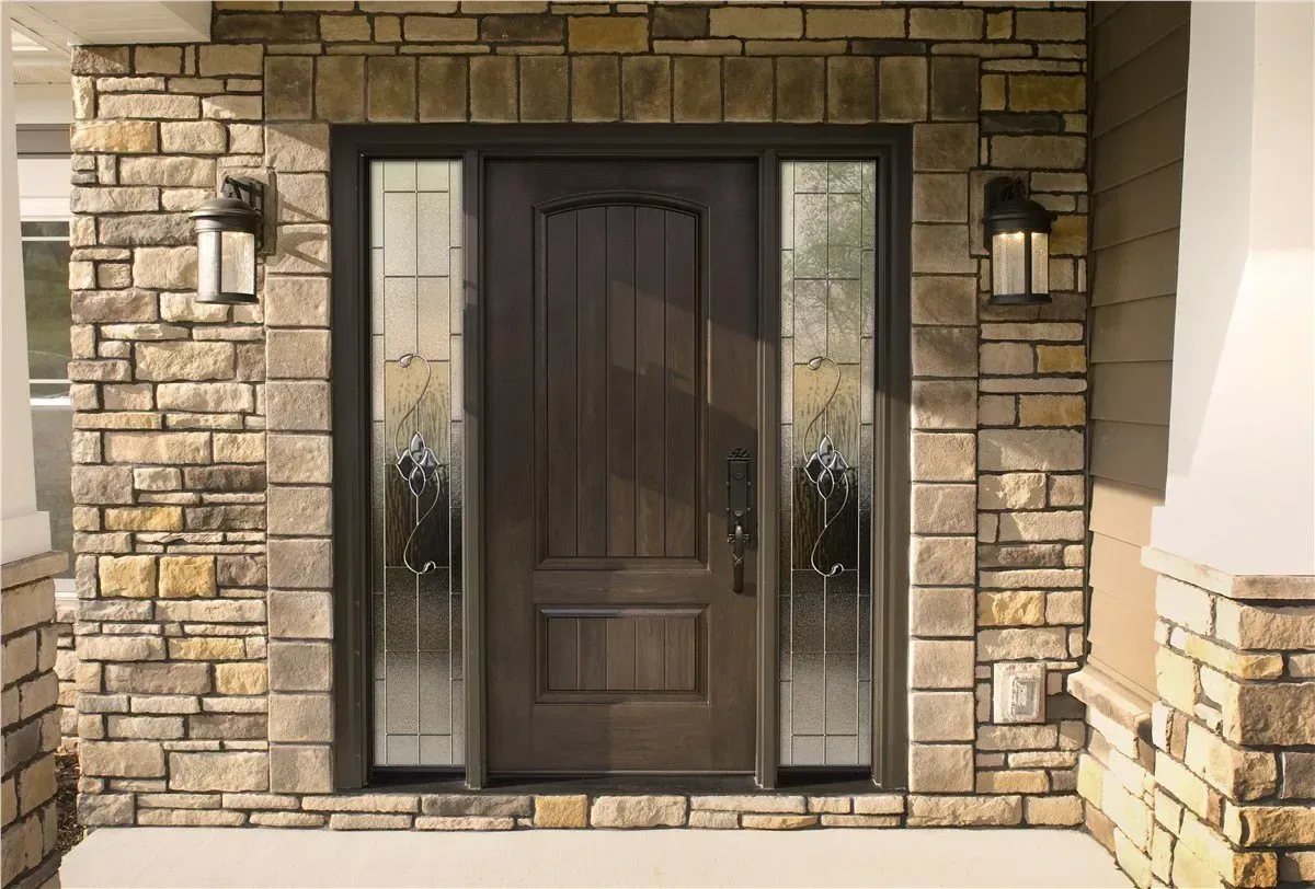 Front entry featuring a dark wood exterior door with vertical panels and an arched top section, flanked by two decorative glass sidelights with ornate swirl patterns. The door is set in a stone facade with warm, natural tones and is framed by matching lantern-style sconces on either side.