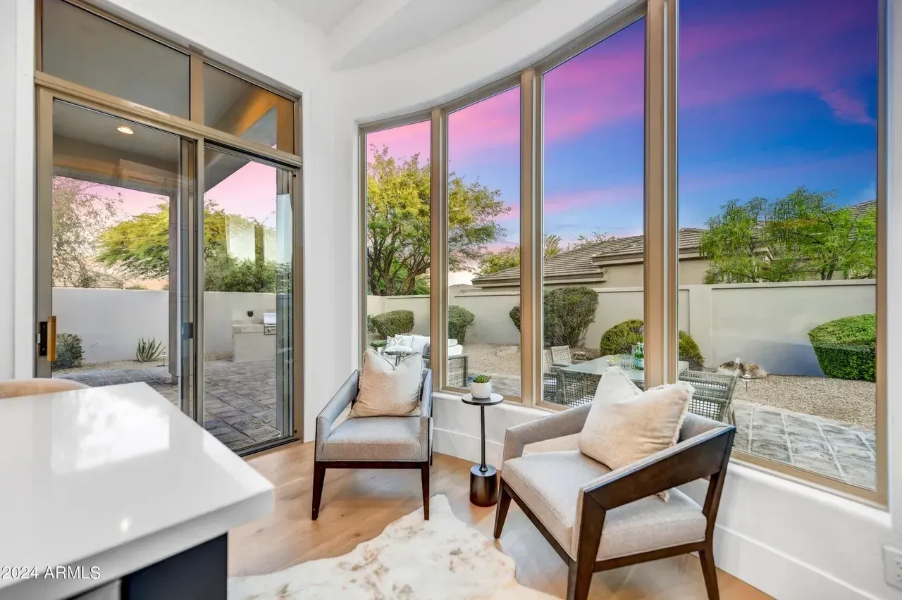 Window installation in a Goodyear home featuring tall, curved floor-to-ceiling glass panels with bronze frames that flood the space with natural light. The windows offer expansive views of a landscaped backyard and a vibrant desert sunset, enhancing the open, airy feel of the cozy indoor seating area with modern chairs and light wood flooring.