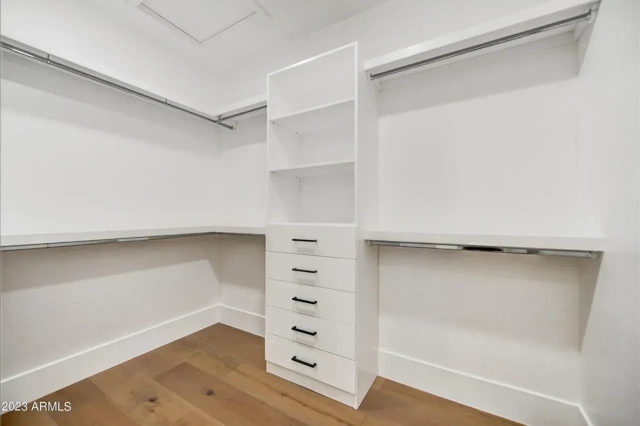 Custom walk-in closet with white built-in shelving and six drawers featuring black handles, surrounded by wraparound hanging rods. The space has white walls, a smooth ceiling with an attic access panel, and wide-plank wood flooring for a clean, organized look.