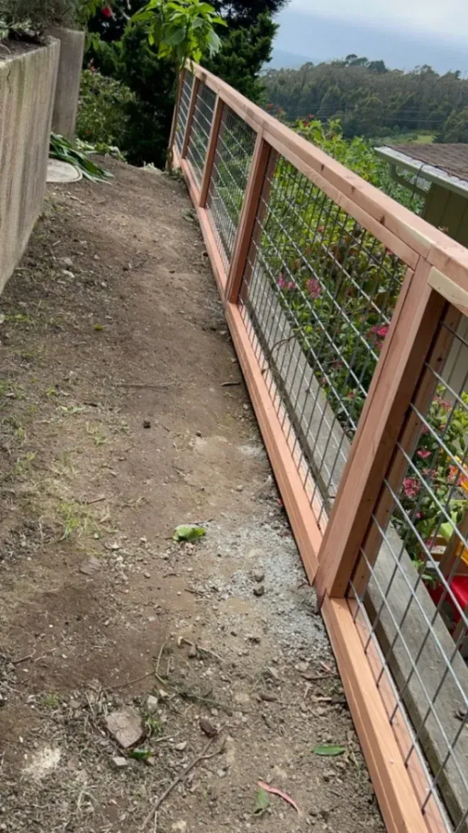 A slightly angled shot of a newly constructed wooden fence with wire mesh panels running along a sloped dirt path.