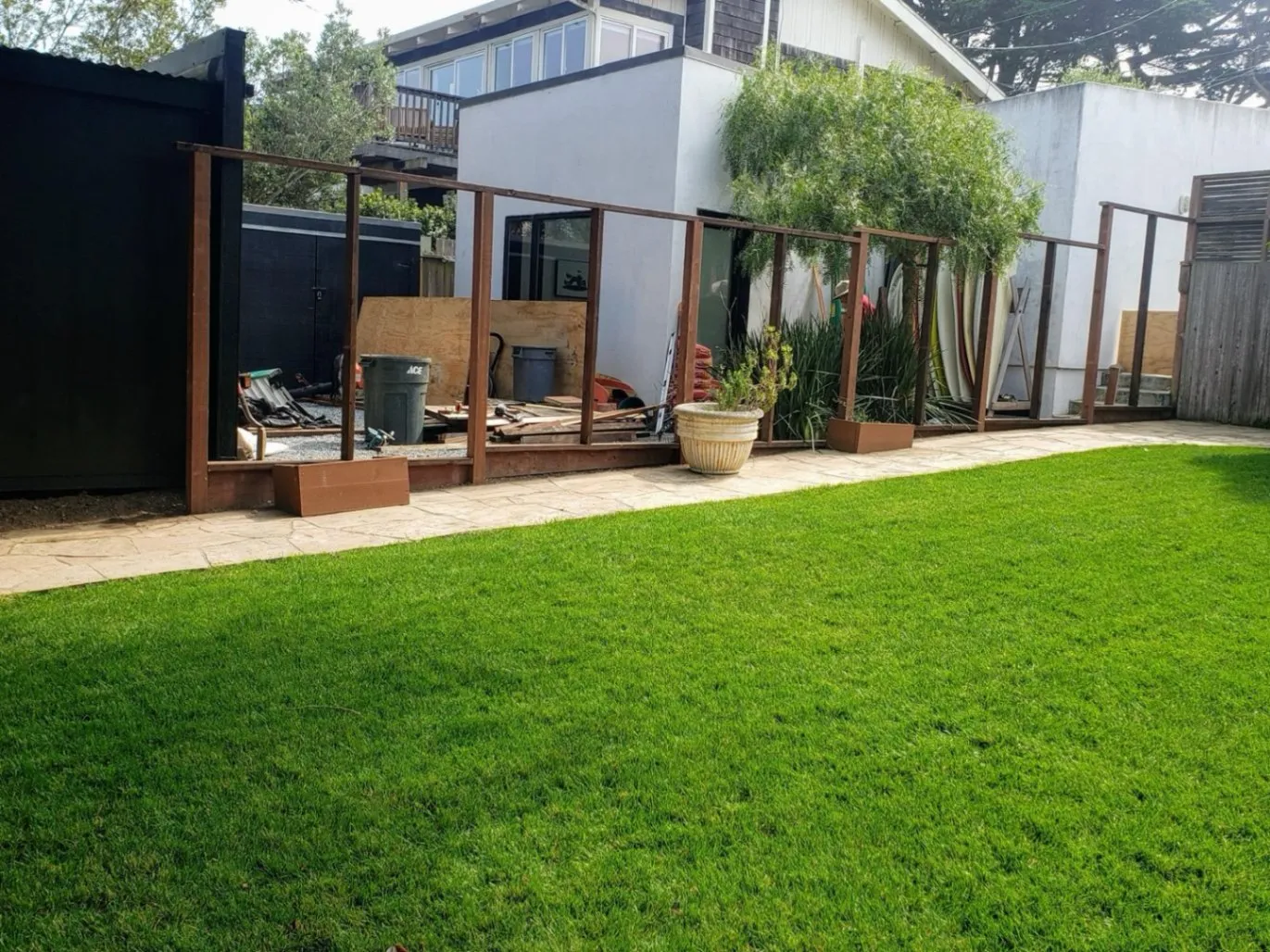Lush green lawn in the foreground, contrasting with a partially built wooden fence and a cluttered backyard under construction.
