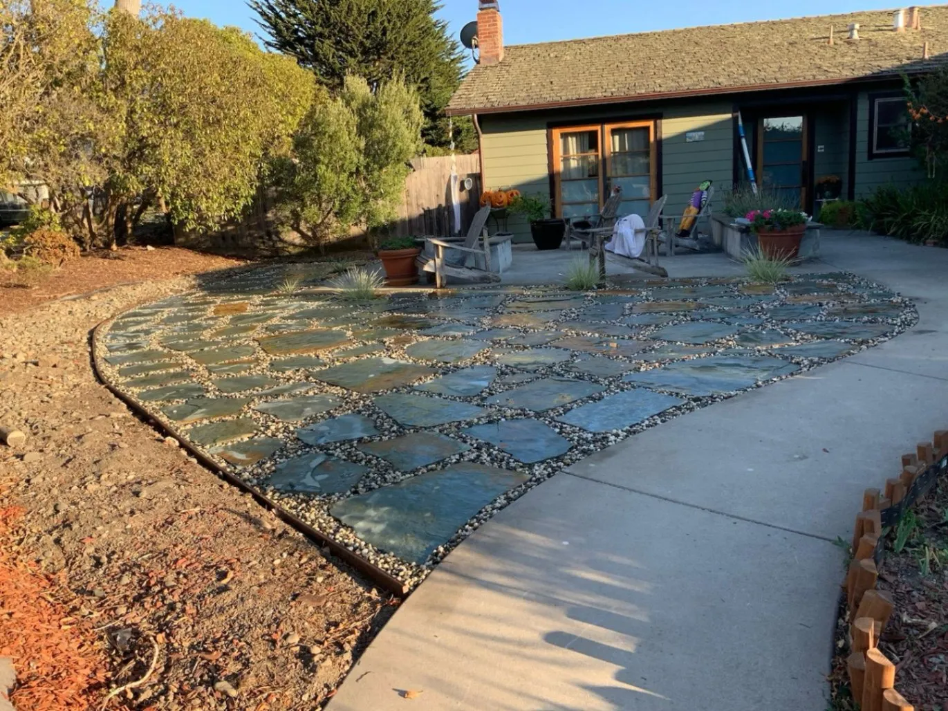 Cate Landscaping created a backyard patio with a unique flagstone and pebble design. The stones are laid in a geometric pattern, surrounded by small pebbles, leading up to a cozy seating area with Adirondack chairs.