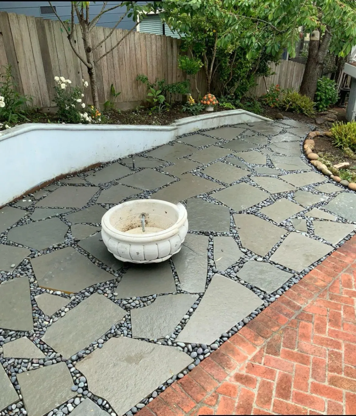 Flagstone patio with dark pebble grout, featuring a small, empty fountain in the center, surrounded by a garden with plants and a wooden fence
