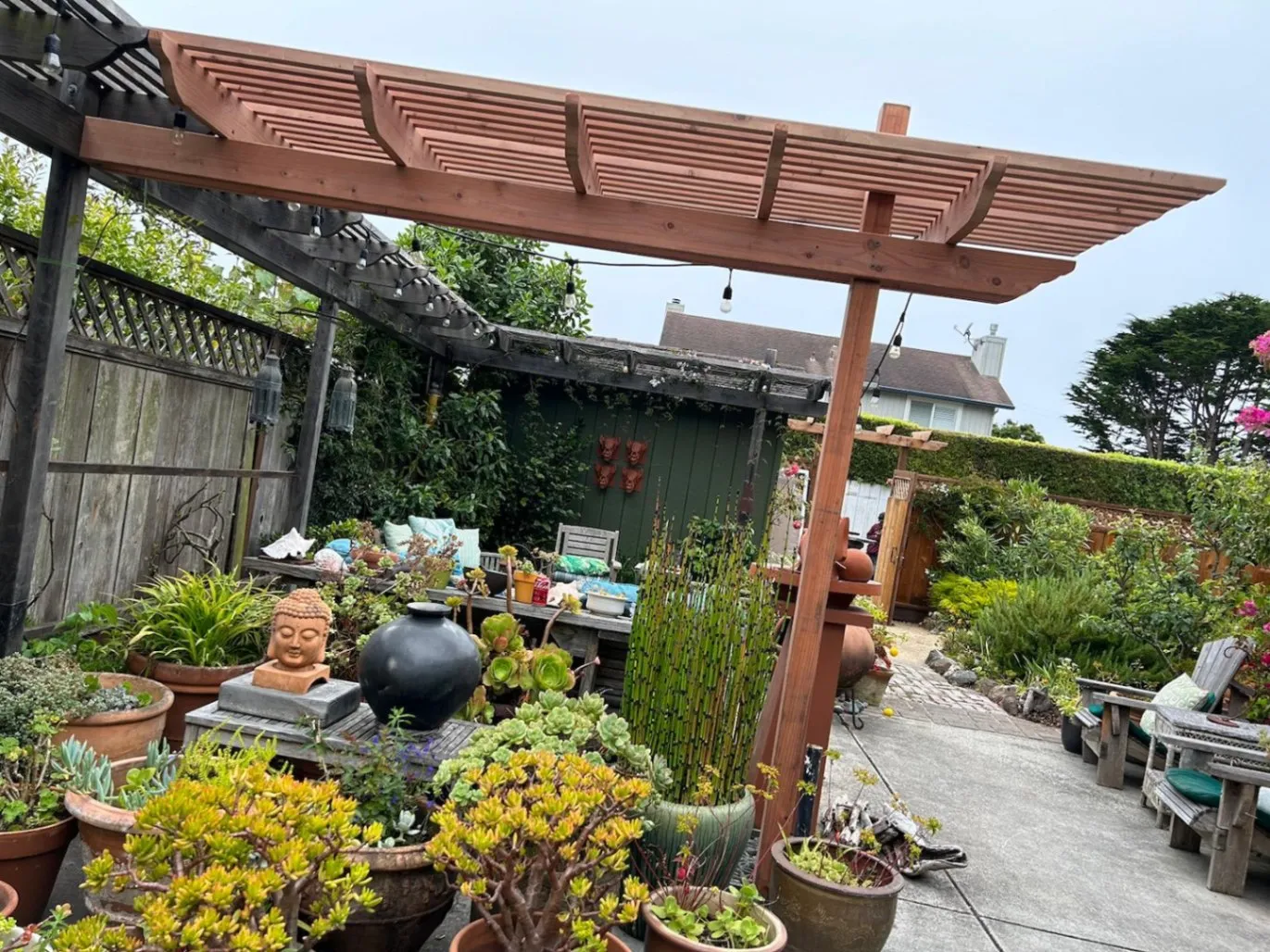 A lush, well-decorated backyard garden with a wooden pergola featuring a slatted roof. The space is filled with potted succulents, greenery, and decorative elements, including a Buddha head sculpture, a black ceramic vase, and hanging lanterns.