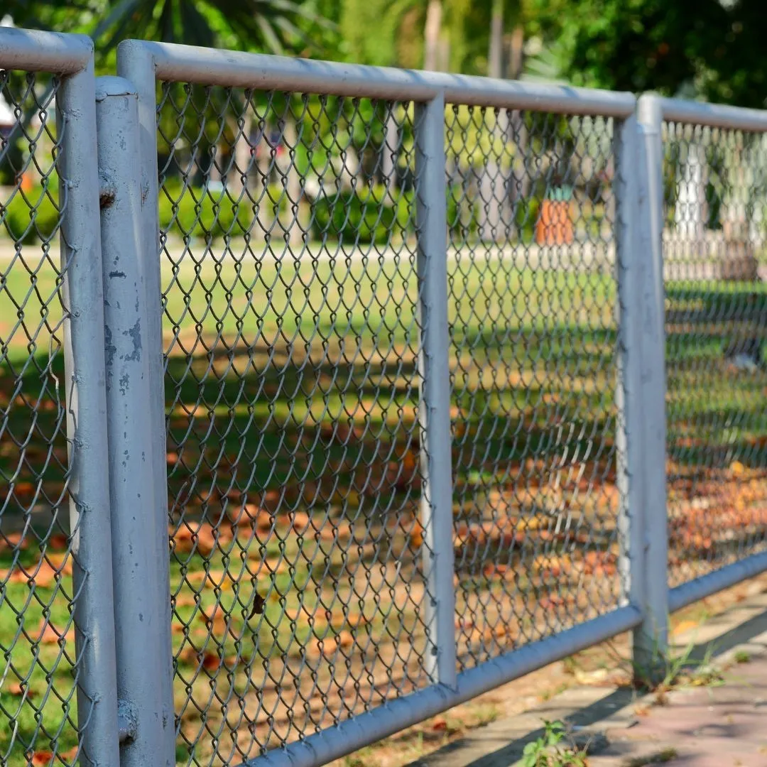 A close-up of a gray metal chain link fence with diamond-shaped mesh and round metal posts, outdoors with greenery in the background.