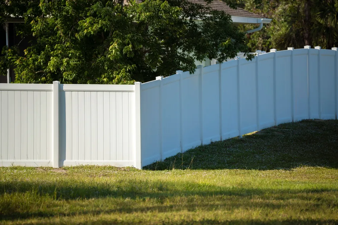 A close-up of a white vinyl privacy fence with a house and trees in the background