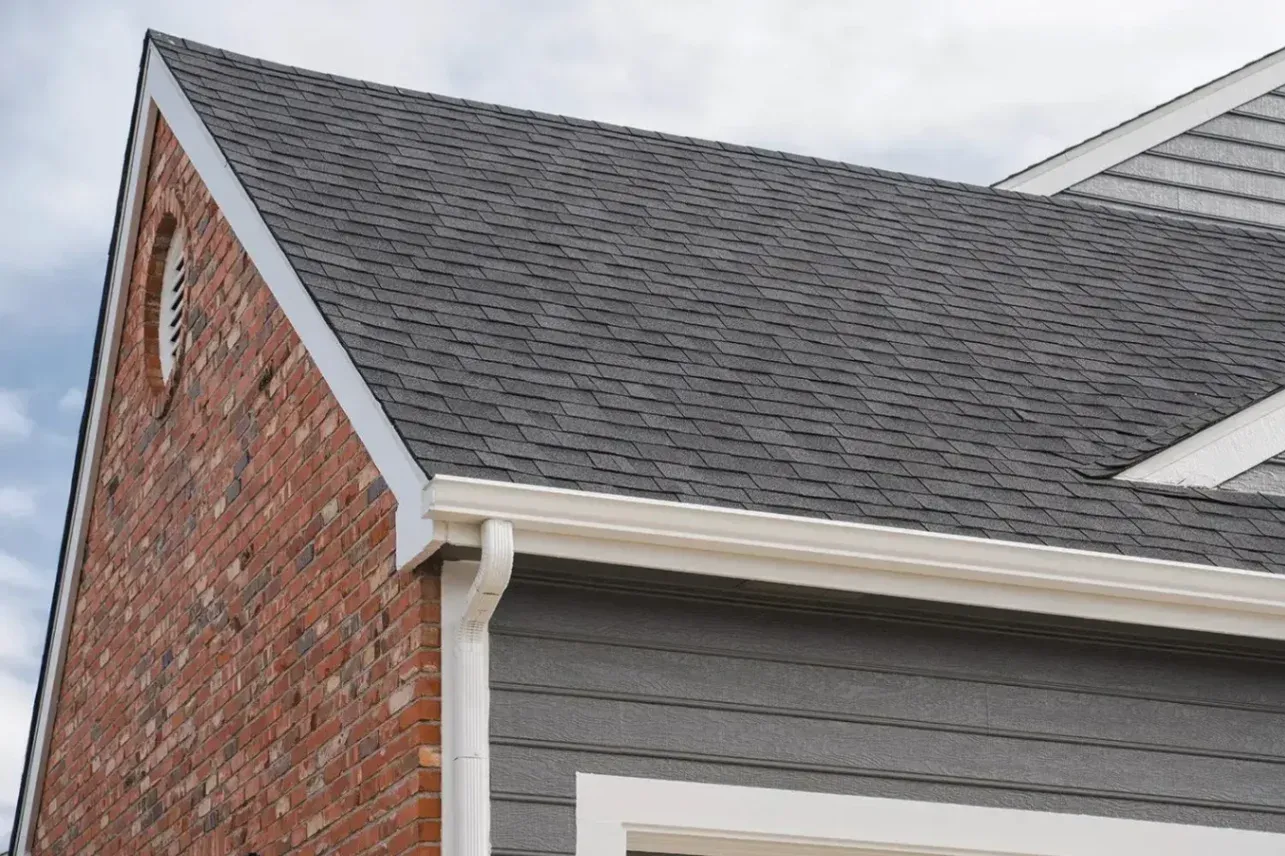 Dark gray architectural asphalt shingles installed on a home roof with white gutters, red brick gable end, and circular attic vent.