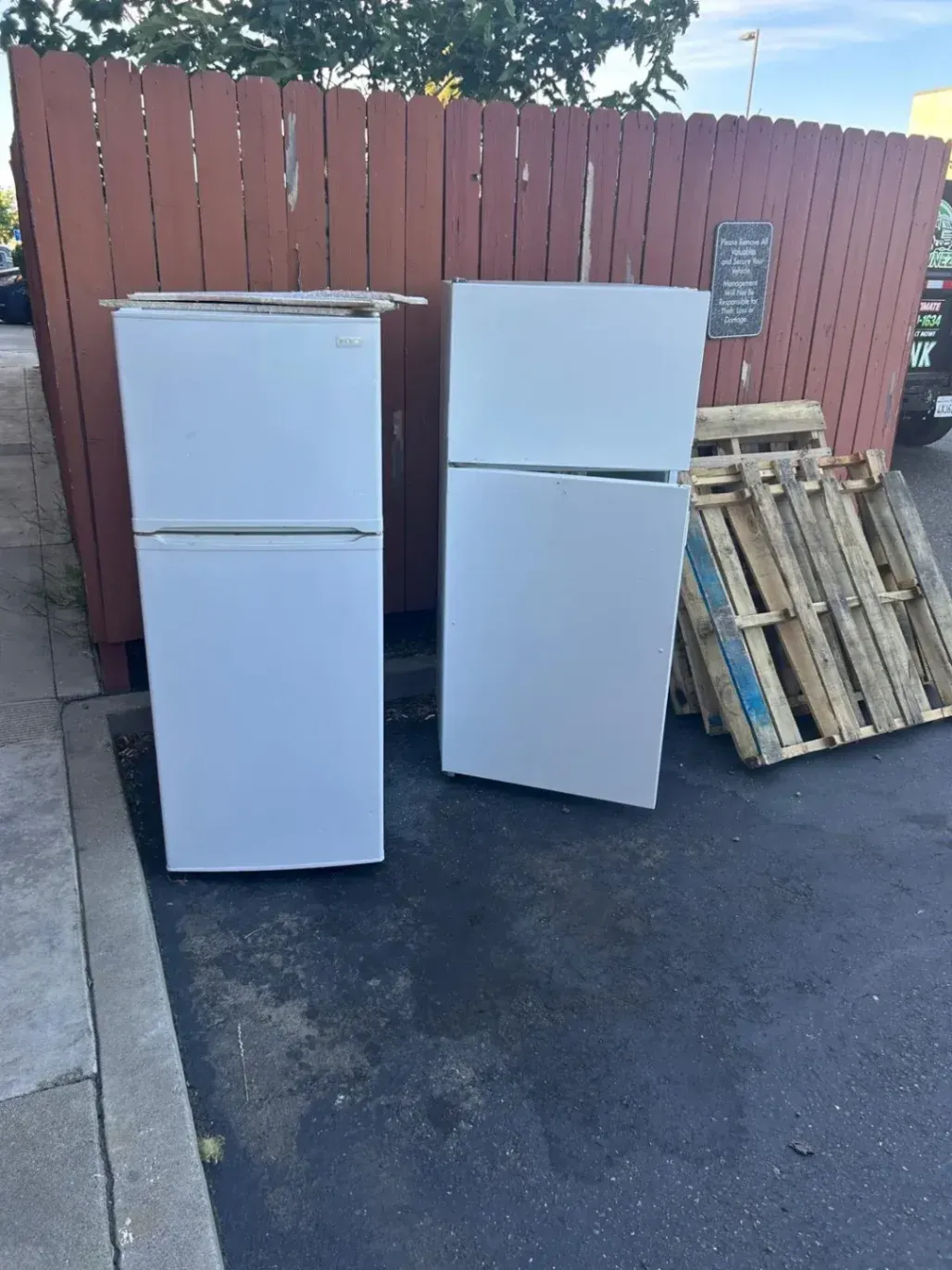 Two old white refrigerators and wooden pallets sitting outside on asphalt next to a wooden fence, ready for appliance removal hauling services in Fairfield, CA.
