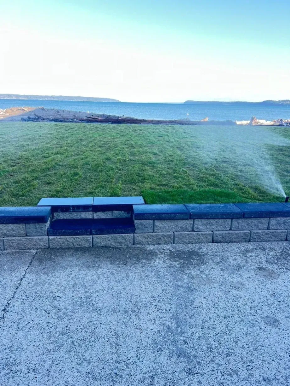 Freshly installed lawn with a stone retaining wall and ocean view in Snohomish, WA, featuring an active sprinkler watering the grass.