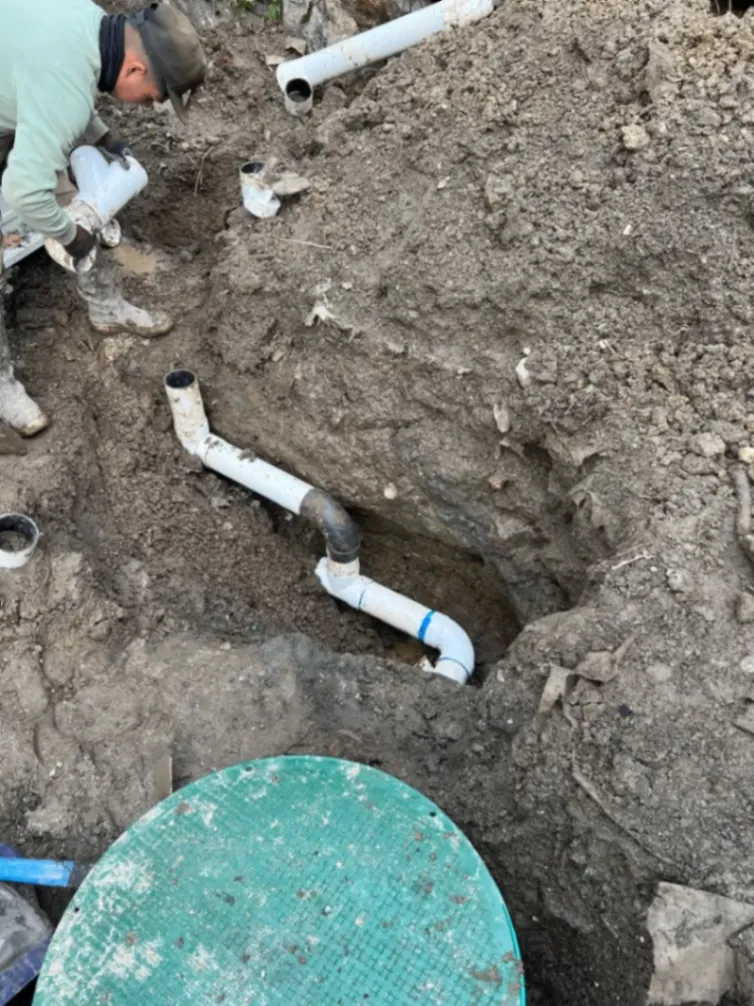 A worker repairing or installing a drainage pipe system in a dug-out hole, surrounded by dirt and plumbing materials.