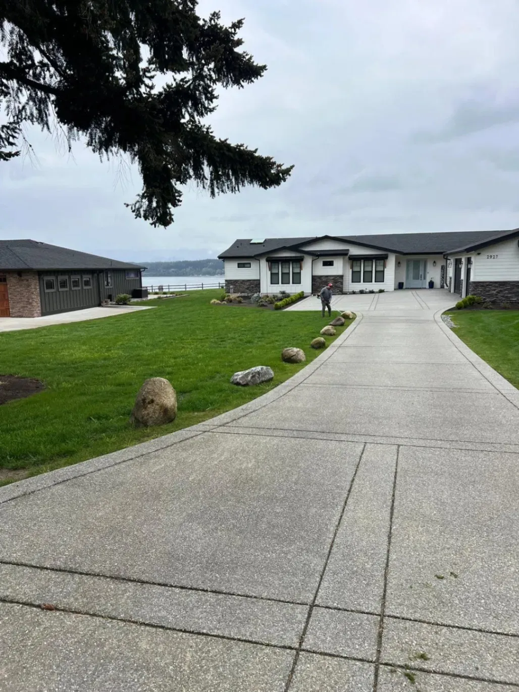 A long, paved driveway leading to a modern house with a neatly manicured lawn and large boulders placed as landscape accents, with a view of a waterfront in the background.